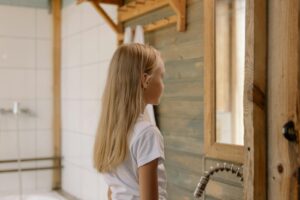 A young girl with blonde hair gazing at her reflection in a rustic wooden bathroom setting.